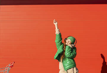 Lächelnde Frau mit drahtlosen Kopfhörern, die Musik hört und vor einer roten Wand tanzt - ADF00221