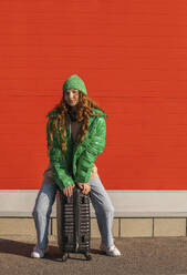 Young woman wearing jacket sitting on suitcase and waiting in front of red wall - ADF00224
