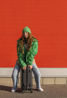 Junge Frau mit Jacke, die auf einem Koffer sitzt und vor einer roten Wand wartet - ADF00224