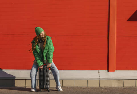 Smiling woman sitting on suitcase and waiting in front of red wall - ADF00225