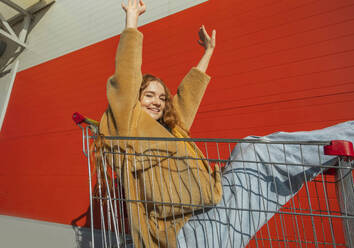 Verspielte Frau mit erhobenen Armen, die in einem Einkaufswagen in der Nähe einer roten Wand sitzt - ADF00231