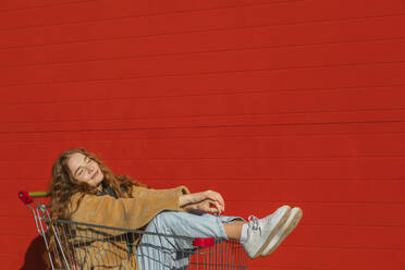Lächelnde Frau mit geschlossenen Augen sitzt in einem Einkaufswagen in der Nähe einer roten Wand - ADF00232