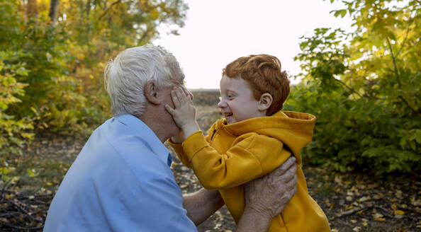 Cheerful boy embracing with grandfather in autumn forest - MBLF00057