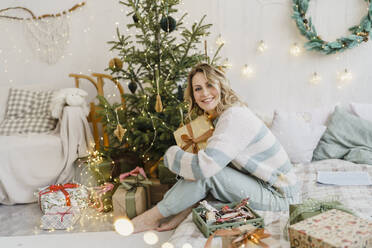 Happy woman holding gift box and sitting near Christmas tree at home - VBUF00465