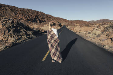 Junge Frau mit Decke geht auf der Straße in der Nähe von Vulkanlandschaft - MMPF00986