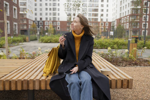 Smiling woman with smart phone sitting on bench and eating lunch at park - LESF00501