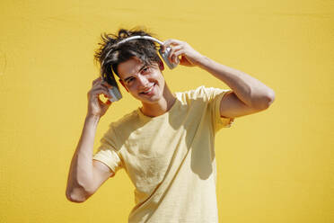 Lächelnder Mann mit drahtlosen Kopfhörern vor einem gelben Hintergrund - NLAF00163