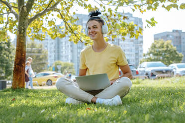 Lächelnder Mann mit drahtlosen Kopfhörern sitzt mit Laptop im Gras - NLAF00167