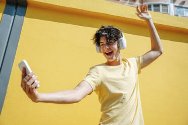 Smiling man wearing wireless headphones taking selfie in front of yellow wall - NLAF00172