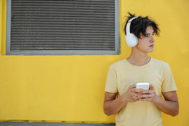 Nachdenklicher Mann mit drahtlosen Kopfhörern und Smartphone vor einer gelben Wand - NLAF00173