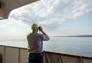 Älterer Mann schaut vom Schiff aus durch ein Fernglas aufs Meer - MBLF00066