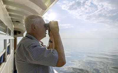 Curious senior man looking at sea through binoculars from ship - MBLF00067