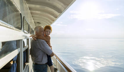 Senior man carrying grandson in arms and looking at sea from ship - MBLF00068