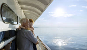 Älterer Mann schaut vom Schiff aus auf das Meer und trägt seinen Enkel an einem sonnigen Tag - MBLF00069