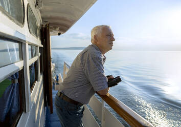 Älterer Mann mit Fernglas in der Hand, der an einem sonnigen Tag vom Schiff aus auf das Meer schaut - MBLF00072