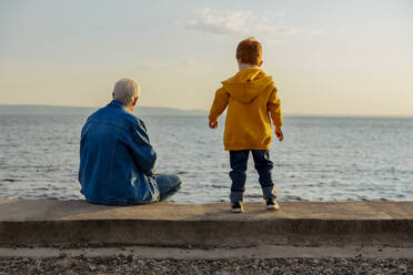 Senior man looking at sea with grandson at sunset - MBLF00082