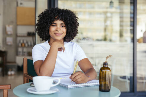 Smiling woman with curly hair writing on note pad at sidewalk cafe - EBSF04014