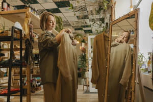 Smiling woman holding dress and standing in front of mirror at store - VIVF01121