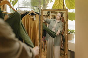 Frau, die in einem Geschäft vor einem Spiegel stehend ein Kleid begutachtet - VIVF01123