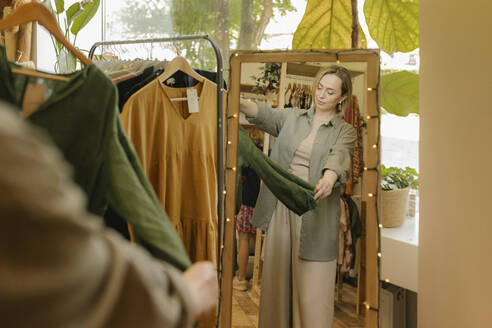 Woman examining dress standing in front of mirror at store - VIVF01123