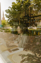 Lächelnder Freiberufler, der in einem Café an einem Laptop arbeitet, gesehen durch ein Glasfenster - VIVF01144