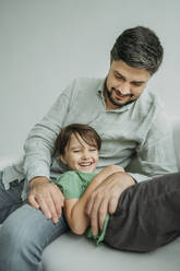 Happy man with son lying on sofa against white background - ANAF02350