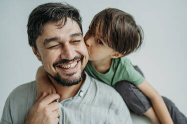 Son kissing happy father on cheeks against white background - ANAF02355