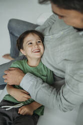 Smiling boy lying on father's lap on sofa - ANAF02358