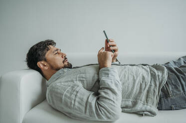 Man using smart phone lying on sofa against white background - ANAF02363