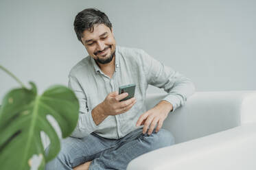 Happy man using smart phone on sofa in front of white wall - ANAF02364