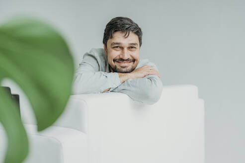 Happy man leaning on sofa in front of white wall - ANAF02366