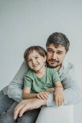 Lächelnder Mann mit Sohn auf Sofa sitzend vor weißem Hintergrund - ANAF02367