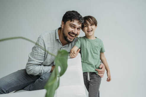 Glücklicher Sohn mit Vater auf Sofa sitzend vor weißem Hintergrund - ANAF02370
