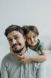 Smiling father sitting on sofa with son in front of white wall - ANAF02375