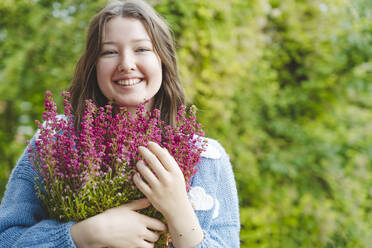 Smiling girl holding heather flowers in front of plants - IHF01773