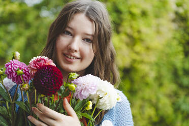 Smiling teenage girl holding dahlia flowers in front of plants - IHF01776