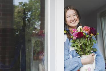 Smiling teenage girl holding dahlia flowers and leaning on window - IHF01778