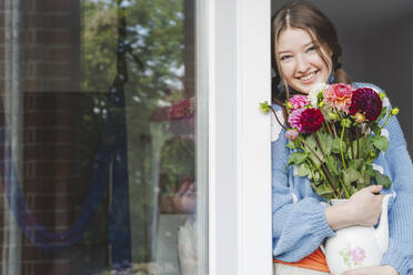 Happy teenage girl holding dahlia flowers and leaning on window - IHF01779