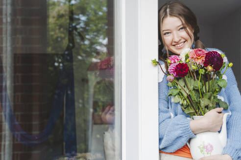 Glückliches Teenager-Mädchen, das Dahlienblüten hält und sich ans Fenster lehnt - IHF01779