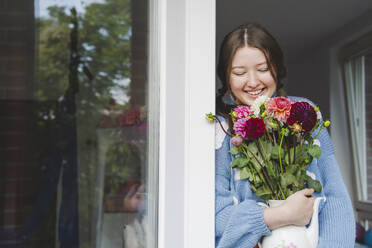 Smiling teenage girl holding various dahlia flowers and leaning on window - IHF01780