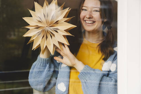 Smiling teenage girl holding star shaped Christmas decoration seen through glass - IHF01785