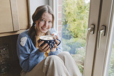 Lächelndes Mädchen sitzt mit heißer Schokolade am Fenster zu Hause - IHF01791