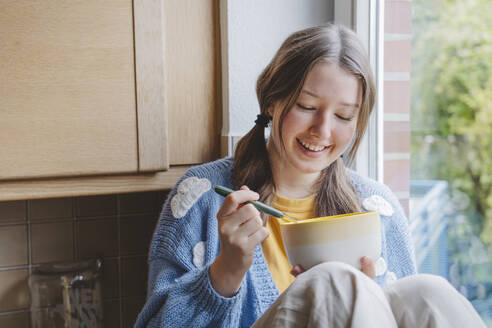 Smiling girl sitting with bowl in kitchen at home - IHF01803