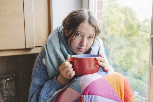 Krankes Mädchen, eingewickelt in eine Decke und sitzend mit einer Kaffeetasse zu Hause - IHF01815