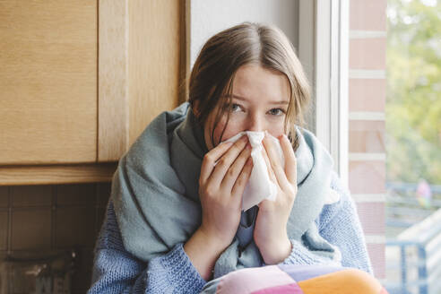 Teenage girl blowing nose on tissue at home - IHF01817