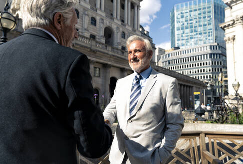 Happy senior businessmen shaking hands together in front of buildings on sunny day - OIPF03573