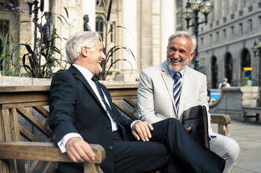 Fröhliche Geschäftsleute sitzen lachend auf einer Bank vor einem Finanzgebäude - OIPF03582