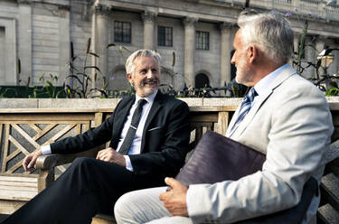 Lächelnde ältere Geschäftsleute, die auf einer Bank vor einem Finanzgebäude miteinander diskutieren - OIPF03584