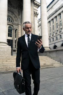 Älterer Geschäftsmann, der in der Nähe eines Gebäudes spazieren geht und sein Smartphone benutzt - OIPF03600