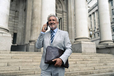 Lächelnder älterer Geschäftsmann, der in der Nähe eines Gebäudes mit einem Smartphone spricht - OIPF03603